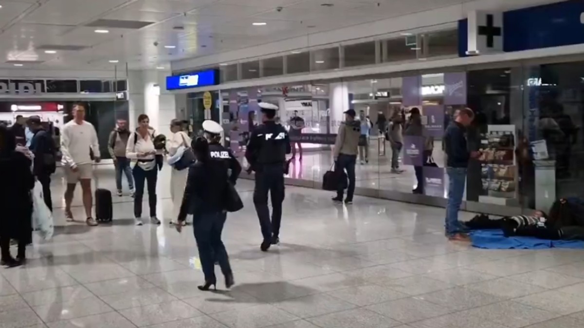 Imaginea surprinde o scenă dintr-un terminal de aeroport aglomerat (posibil Aeroportul München, conform numelui fișierului), unde pasagerii circulă printre magazine. În centrul scenei, doi ofițeri de poliție (cu inscripția "POLIZEI" pe spate) patrulează, în timp ce un om este întins pe jos, pe o saltea, lângă vitrinele magazinelor.
