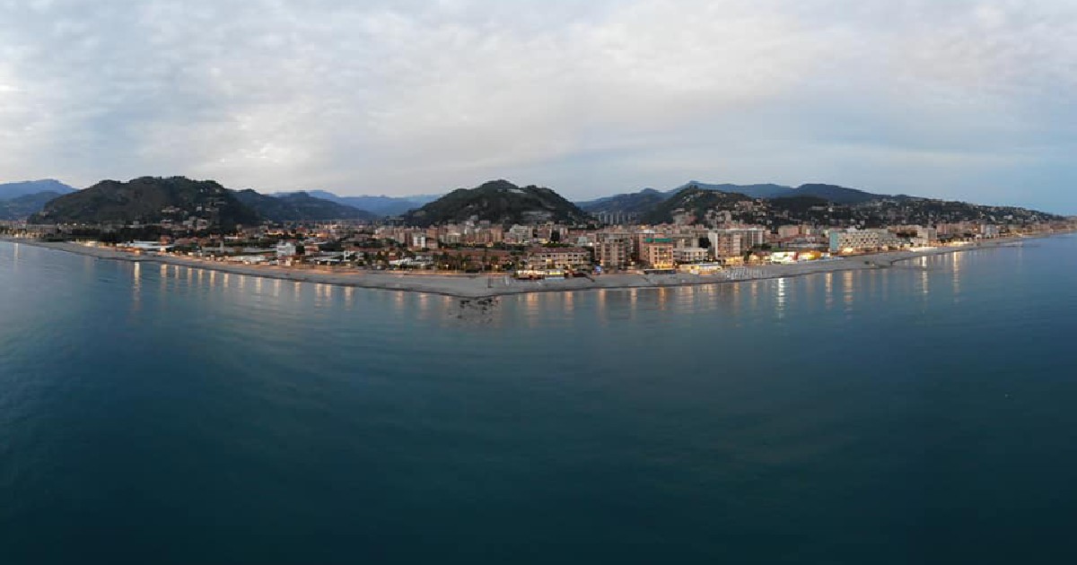 O fotografie panoramică de la înălțime surprinde o localitate de coastă iluminată la amurg, întinsă de-a lungul unei plaje curbate, cu munți în fundal.