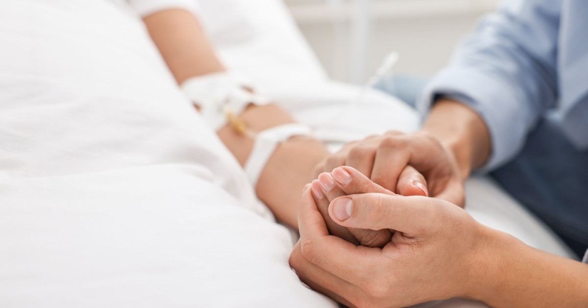 Imagine cu mana unui pacient, pe patul de spital, tinuta de ambele maini ale unei persoane dragi