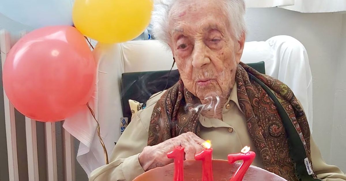Maria Branyas Morera, pe scaun, cu un tort cu lumânări aprinse de culoare roșie care formează numărul 117, cu baloane roșii și galbene pe fundal