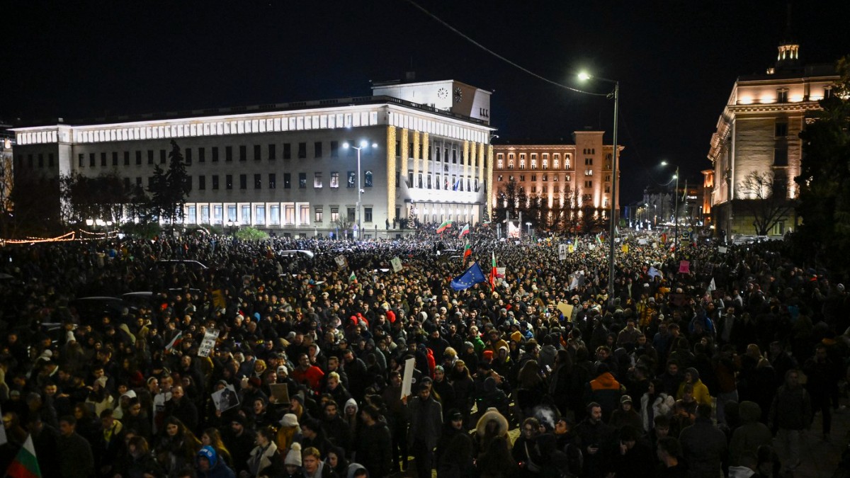Imaginea arată o mulțime uriașă de protestatari adunați noaptea într-o piață din centrul unui oraș, fluturând steaguri și pancarte. Clădirile instituțiilor publice din fundal sunt iluminate, evidențiind amploarea și intensitatea manifestației.