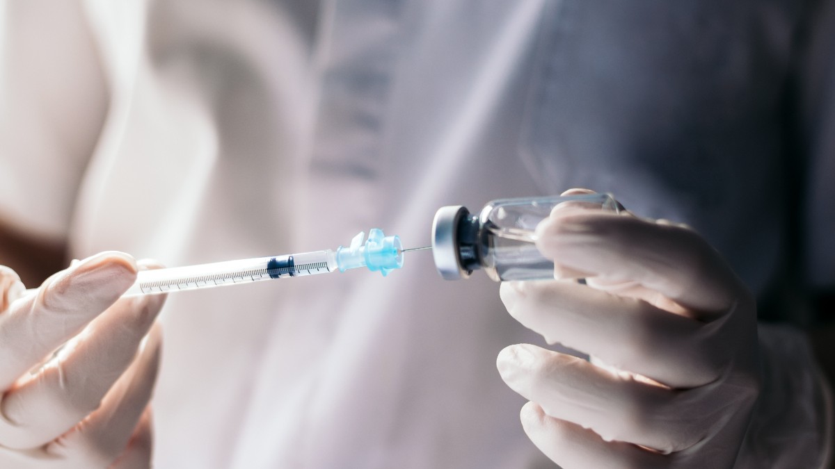 Un cercetător îmbrăcat în echipament medical de protecție pregătește o doză de vaccin, extrăgând lichidul dintr-un flacon cu ajutorul unei seringi. Imaginea sugerează un context de laborator sau de testare clinică, asociat dezvoltării și administrării vaccinurilor.