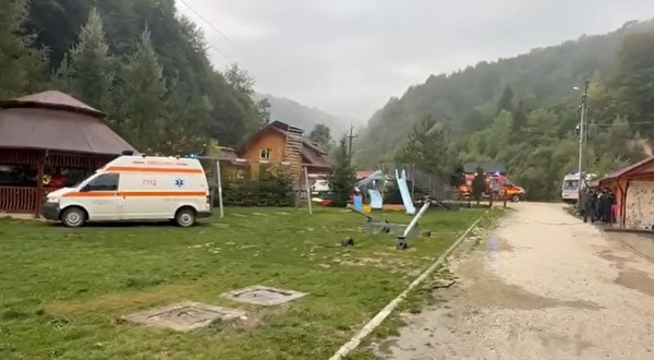 O ambulanță și alte vehicule de intervenție staționate în curtea unei cabane, înconjurată de munți
