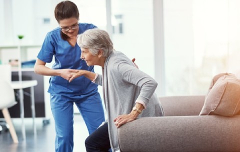 O asistentă medicală o ajută pe o femeie în vârstă să se ridice de pe o canapea gri