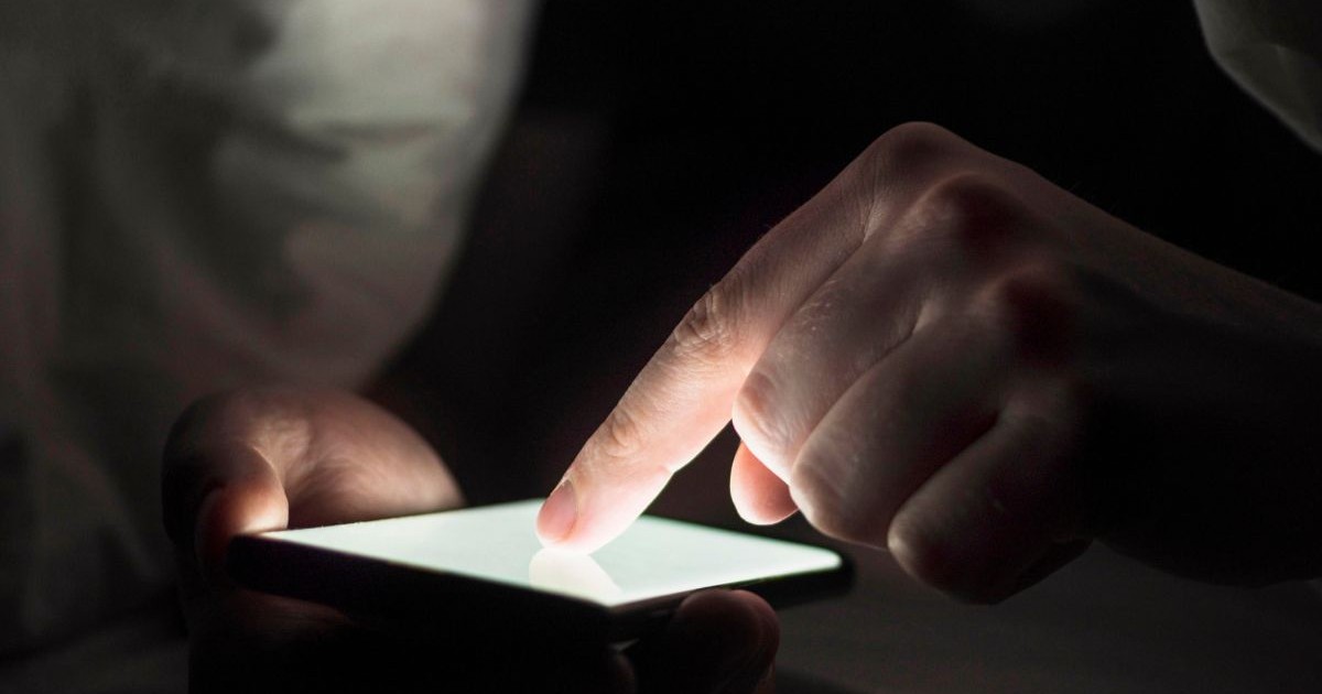 Bărbat care stă pe telefon, ținând degetul arătător lipit de ecranul telefonului mobil care este luminat în alb