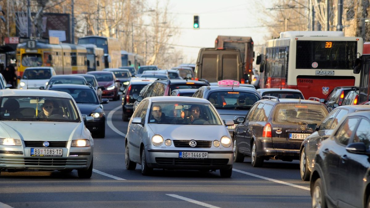 Scena aglomerată a unei străzi urbane în timpul zilei, plină de mașini, autobuze și camioane. Drumul este aglomerat cu vehicule de diferite culori, iar în fundal se văd copaci goi și un semafor.