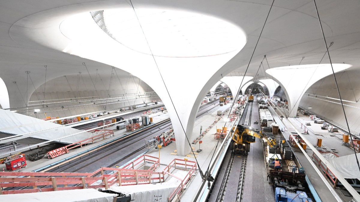 În imagine este prezentat șantierul unei gări subterane moderne cu o arhitectură futuristă din stâlpi albi de beton în formă de potir, linii de cale ferată în curs de amenajare și diverse utilaje de construcție.