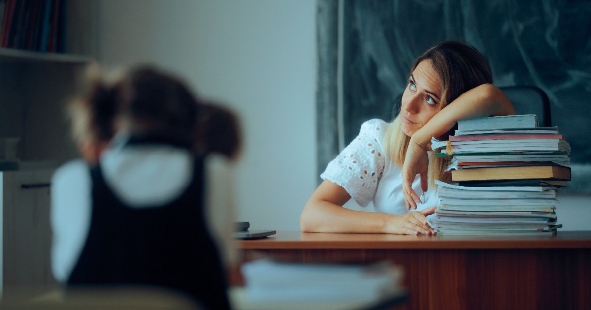 O profesoară la catedră, este supărată și stă cu capul rezemat pe un teanc de cărți și privește în gol