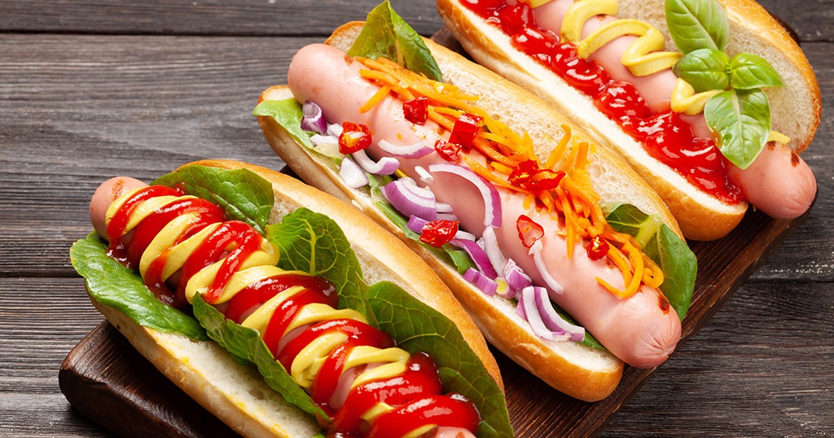 Trei hot-dogi cu sosuri diferite așezate pe masă.