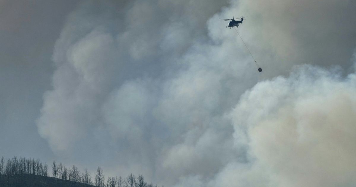 Elicopter pentru stingerea incendiilor, care poartă agățat un obiect rotund, suspendat de o coardă lungă. O mare parte din imagine este ocupată de nori groși de fum, de nuanțe gri. În partea de jos, pe orizontală, apare o colină sau un deal cu vârful marcat de contururile subțiri ale unor copaci fără frunze, rari și răsfirați.