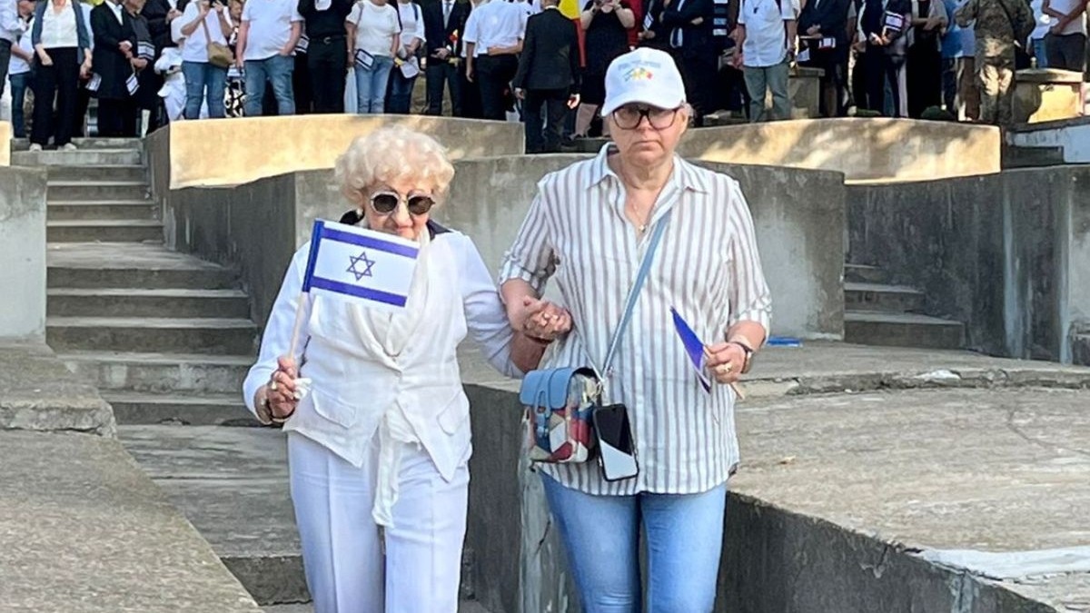 În imagine Rasela Koka Palmon, stanga, supraviețuitoare a Holocaustului la Iași, in varsta de 91 de ani, in haine deschise la culoare, cu un steag mic in mana, la brat cu o femeie cu sapca alba pe cap, merg printre mormitele, in spate se vad multi oameni