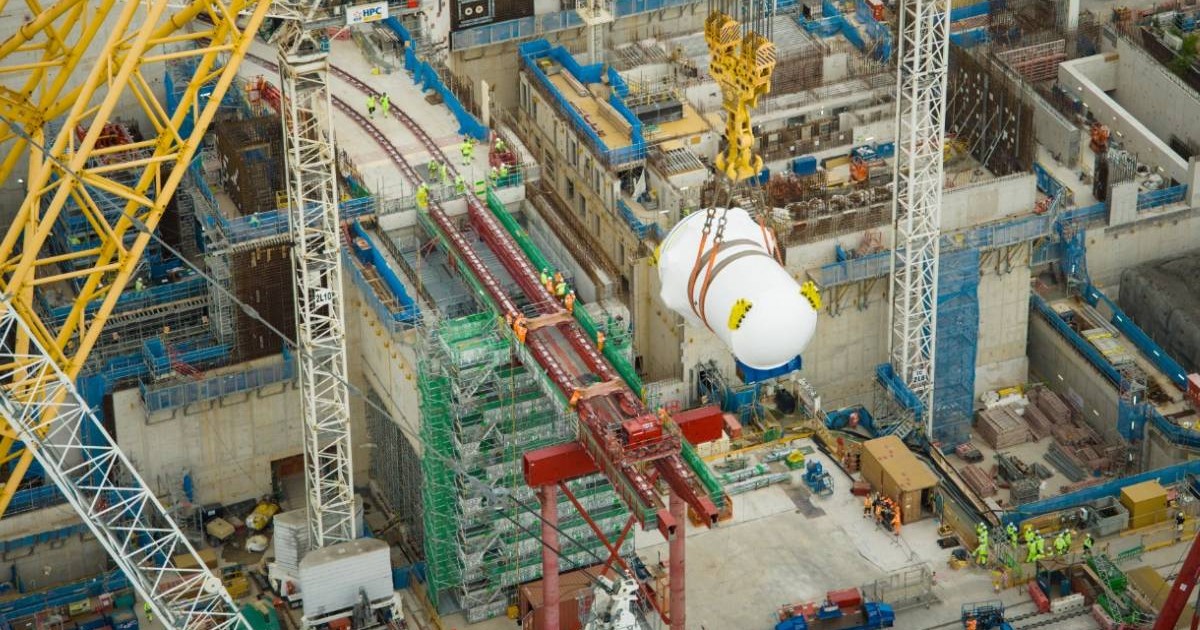 Imagine de pe șantierul centralei nucleare Hinkley Point C din Marea Britanie. Cea mai mare macara din lume ridică un vas alb, uriaș, de reactor nuclear, suspendat de cabluri groase, deasupra unei zone de construcție pline de schele, macarale și muncitori.