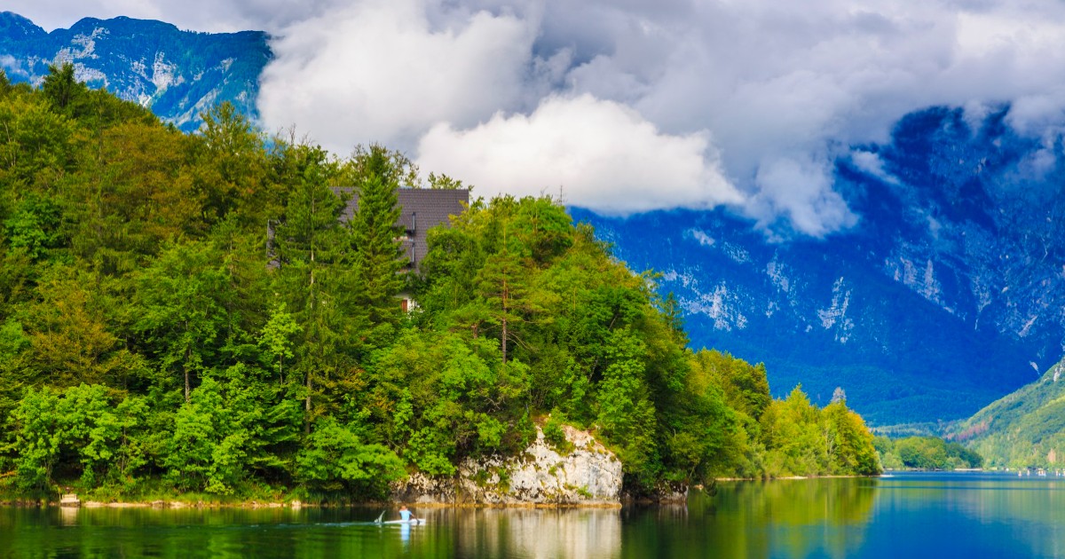 Imaginea surprinde un peisaj montan spectaculos, cu un lac limpede în prim-plan. Pe malul lacului se vede o cabană cu acoperiș închis la culoare.