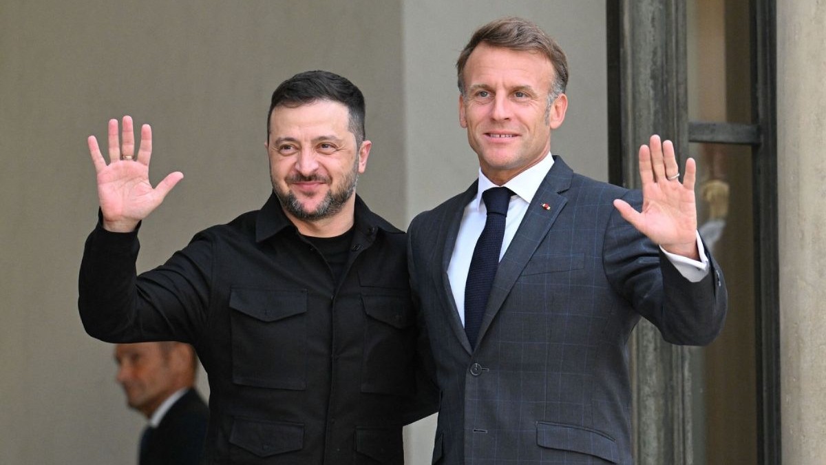 Volodimir Zelenski, în stânga, și Emmanuel Macron, în dreapta, ambii cu mâinile stângi ridicate, în timp ce se țin de mijloc