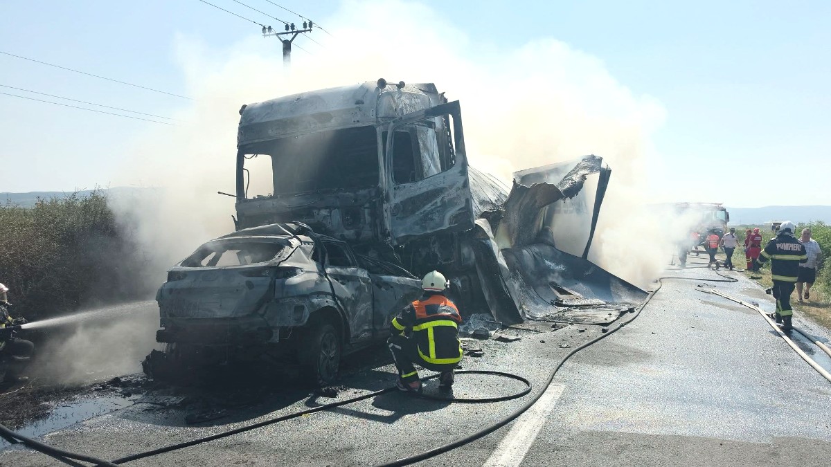 Imaginea prezintă pompierii intervenind la locul unui accident rutier grav, unde un autoturism și un camion au fost complet distruse de un incendiu puternic pe o șosea.