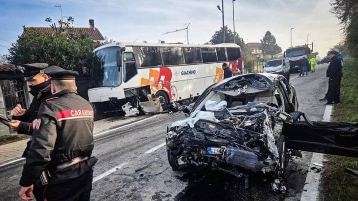 In imaginea se vad doi carabinieri in stanga, in preapta o masina BMW distrusa complet, iar in spate un autocare cu alb, rosu si portocali lovit in partea laterala
