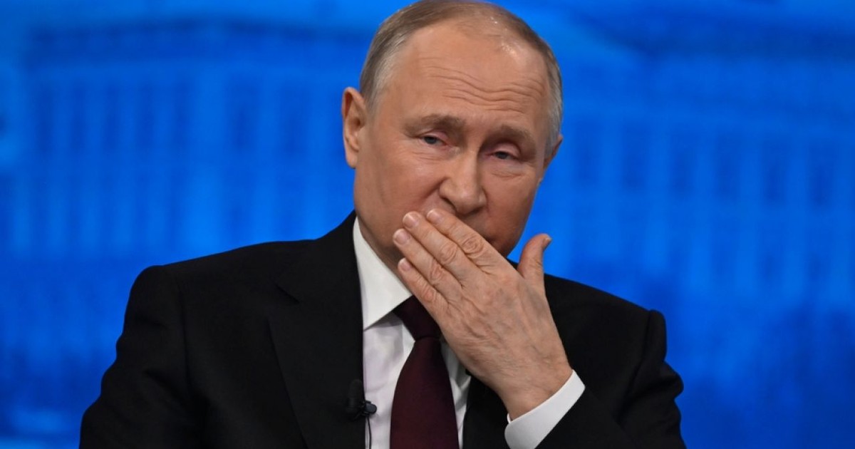 Vladimir Putin, îmbrăcat în costum negru, cu cămașă albă și cravată albastră, cu mâna la gură, pe fundal de culoare albastră
