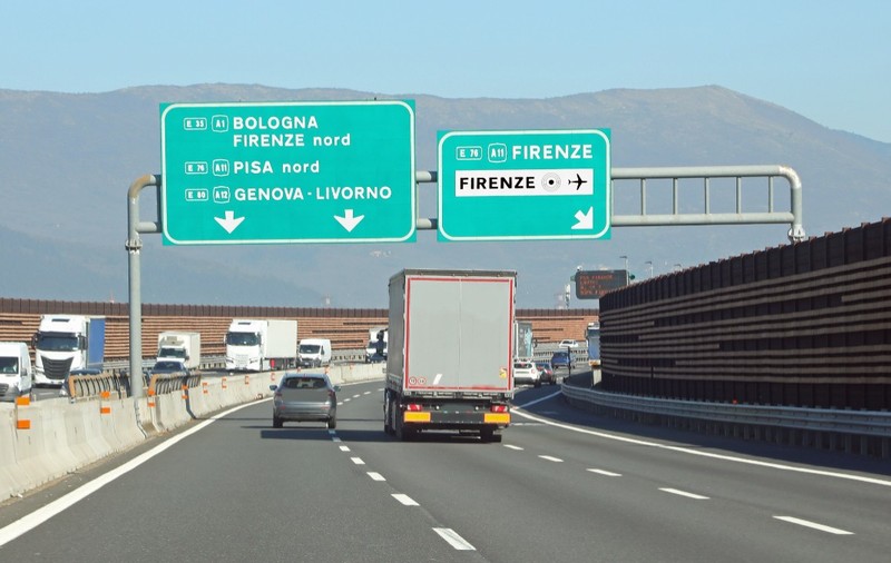 Traficul de pe o autostradă din Italia, cu mai multe autovehicule și camioane deplasându-se pe benzi sub panouri verzi mari de semnalizare rutieră ce indică direcțiile spre Bologna, Firenze, Pisa, Genova și Livorno.