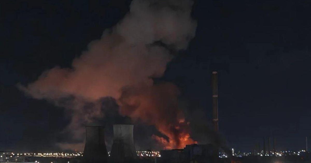 Imaginea surprinde un incendiu puternic izbucnit într-o zonă industrială, cel mai probabil la o centrală termoelectrică, cu flăcări intense și un nor dens de fum ridicându-se în noapte. În fundal se pot observa instalațiile și coșurile industriale, iluminate de luminile orașului, în timp ce focul domină scena.
