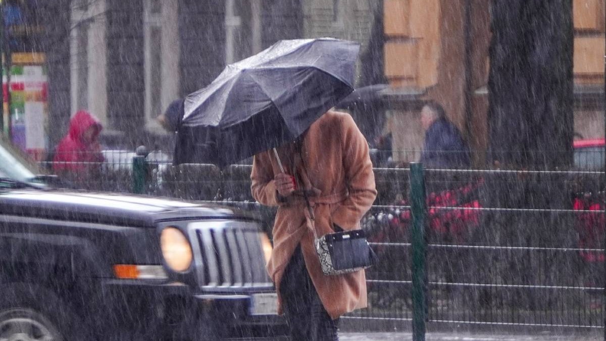 Imaginea surprinde o persoană văzută din fata, cu un palton crem, care se protejează de vant si ploaie sub o umbrelă neagra, într-un cadru urban, se vede o masina neagra cu farurile aprinse, mai multe persoane cu pelerine de ploaie in spate, ploua torential