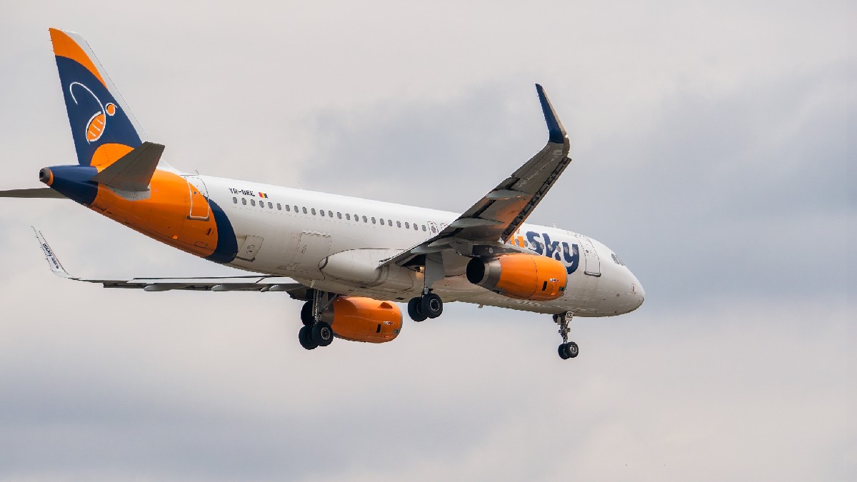 Imaginea prezintă un avion de pasageri în zbor, cu trenul de aterizare coborât. Avionul este vopsit în culorile companiei HiSky, având fuselajul alb, iar motoarele, aripile și coada în nuanțe de portocaliu și albastru. Pe motoare și pe fuselaj este scris numele companiei, "sky". Aeronava este fotografiată pe un cer parțial înnorat.