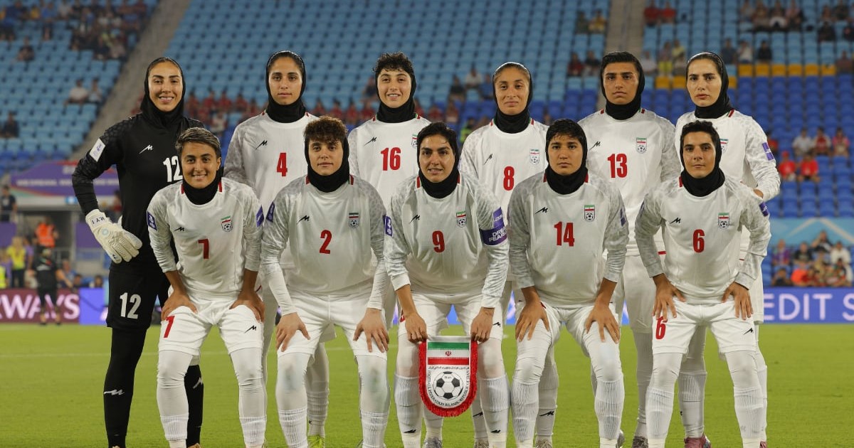 Echipa națională feminină de fotbal a Iranului pozează pe teren înaintea unui meci, aliniată pentru fotografia oficială. Jucătoarele poartă echipament alb și hijab, iar în fundal se pot observa tribunele stadionului.