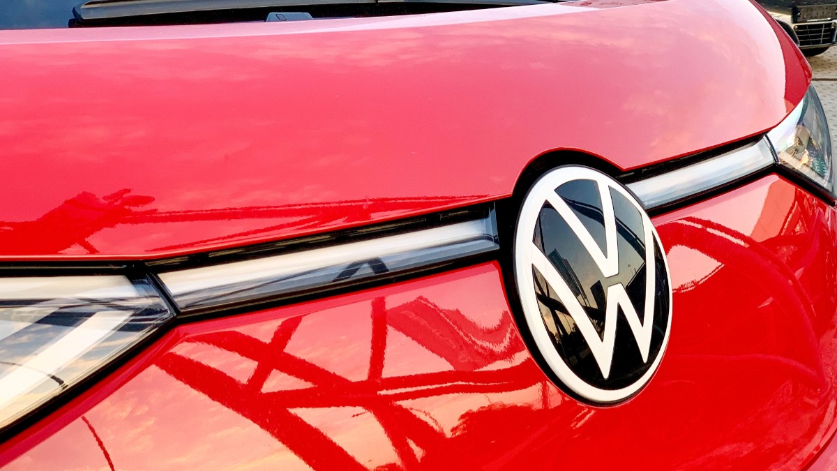 Imaginea prezintă un plan apropiat cu partea frontală a unui autovehicul roșu, evidențiind emblema circulară alb-negru a mărcii Volkswagen