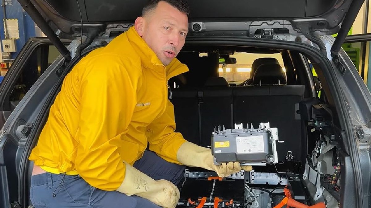 În imagine se vede un bărbat îmbrăcat într-o jachetă galbenă, lângă un BMW X5 plug-in hybrid. Capota mașinii este ridicată, iar în partea din față se observă echipamente, cabluri și scule de lucru, specifice unei reparații sau intervenții tehnice