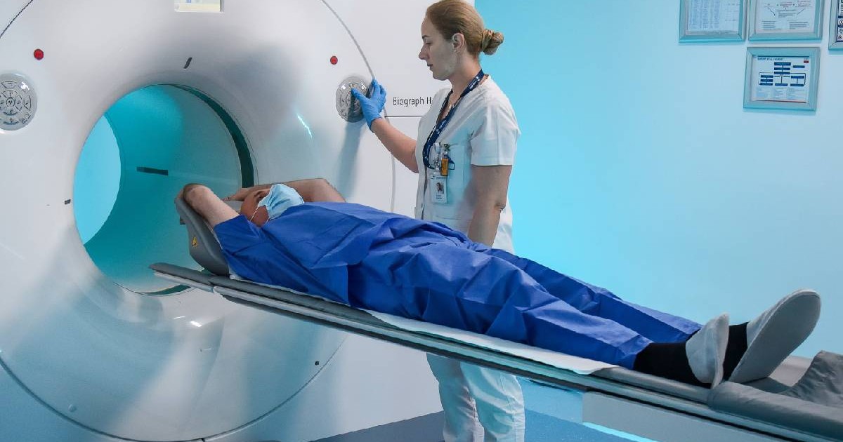 Un pacient imbracat in costum de spital, asezat pe un aparat pentru investigatie si o femeie imbracata in halat alb, cadru medical care seteaza aparatul PET-CT PSMA. Cancer de prostata tehnologie in premiera Sanaddor. Foto: Sanador