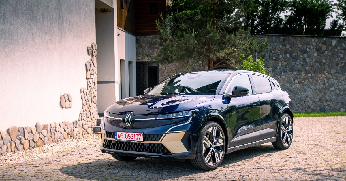 O mașină Renault Megane E-Tech de culoare albastru închis, cu accente aurii pe bara de protecție frontală, este parcată pe o alee pavată cu piatră cubică. Mașina este amplasată în fața unei clădiri cu o fațadă parțial acoperită cu piatră brută și înconjurată de copaci.