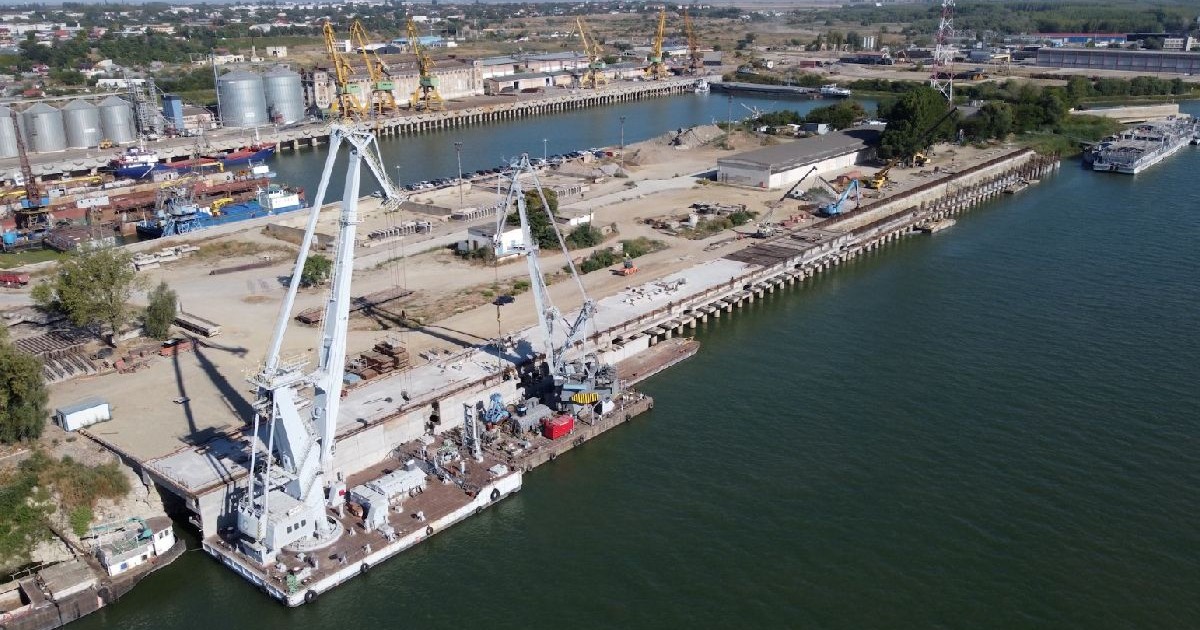 Imaginea prezintă o vedere aeriană a Portului Brăila, unde mai multe macarale de mari dimensiuni sunt poziționate de-a lungul cheiului pentru operațiuni de încărcare și descărcare a navelor.