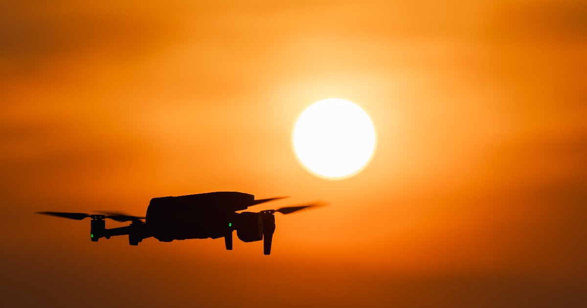 Imaginea surprinde silueta unei drone aflate în zbor pe fundalul unui apus spectaculos, cu soarele dominând cerul în nuanțe intense de portocaliu. Fotografia ilustrează tehnologia modernă a dronelor și utilizarea acestora în spații deschise, fiind potrivită pentru articole despre aviație, inovație sau noile profesii din domeniu.