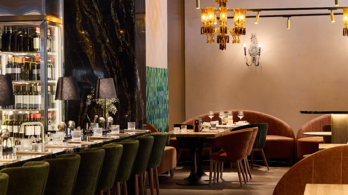 Imaginea prezintă interiorul unui restaurant elegant, cu mese aranjate atent, scaune tapițate în tonuri calde și un decor rafinat, dominat de lumini ambientale și candelabre decorative. Atmosfera sugerează un local de fine dining, cu accent pe confort, intimitate și experiență culinară exclusivistă.