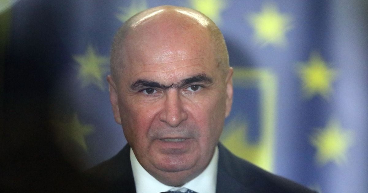 Ilie Bolojan, îmbrăcat în costum negru și cămașă albă, cu logo-ul PNL în blur, pe fundal, și o umbră neagră în partea stângă a fotografiei