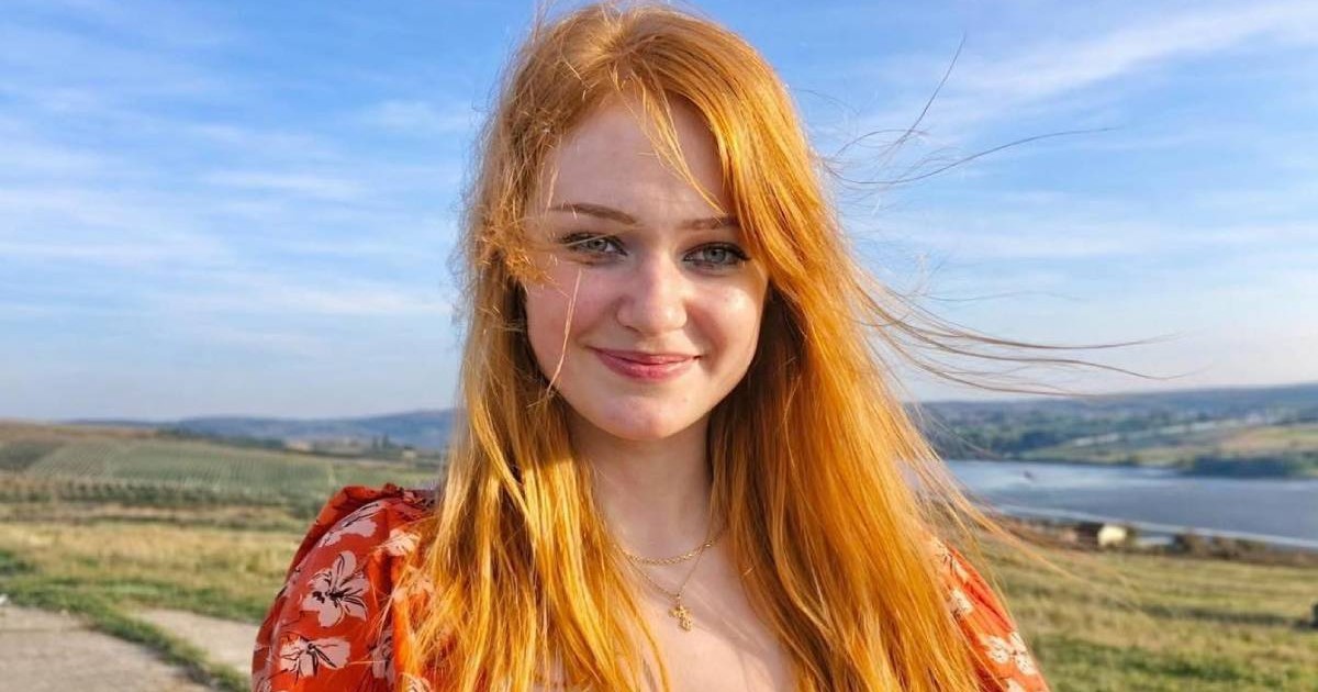 Andreea Anita, cu parul portocaliu, desfacut, zburat de vant, intr-o rochie rosie cu flori albe, zambeste intr-o zi insorita, se vede un cer albastru, in spate dealuri verzi și o apa, Andreea, din Falticeni, are nevoie de 2.3 milioane de dolari pt un tratament in SUA