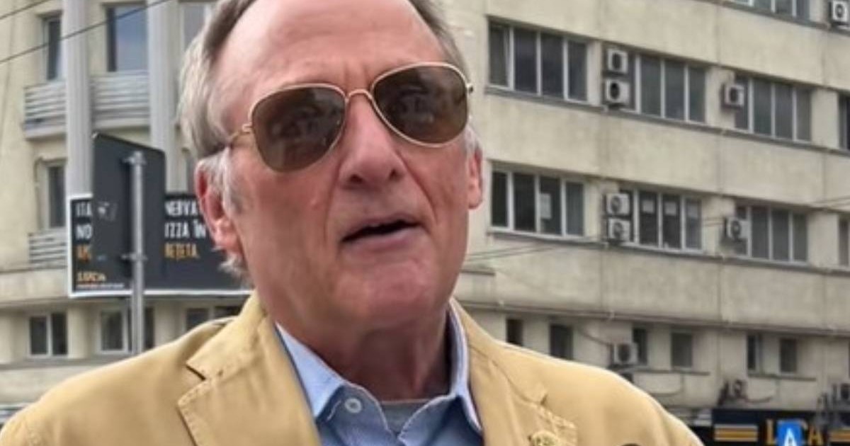 Un bărbat în vârstă, american, îmbrăcat cu un palton bej și o cămașa albastră, în timp ce acordă un interviu, n fundal se vede un bloc din centrul Bucureștiului
