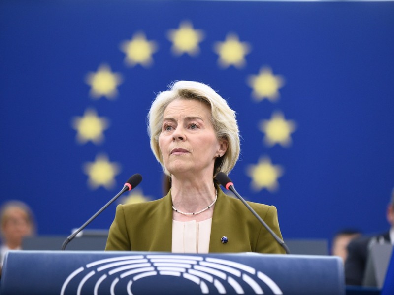 Imaginea o arată pe Ursula von der Leyen vorbind la un pupitru. Are părul scurt, blond și poartă un sacou verde-măsliniu peste un top de culoare crem. Un colier de perle este vizibil la gât. Fundalul este un steag albastru cu un cerc de stele galbene. Două microfoane se află în fața ei Foto: Profimedia