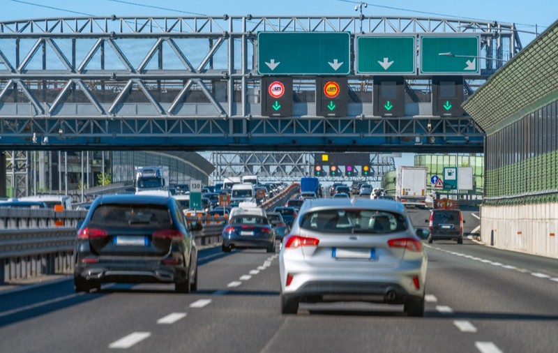 Trafic intens de vehicule pe o autostradă aglomerată, cu mai multe benzi circulabile și panouri electronice de semnalizare montate pe o structură metalică suspendată.