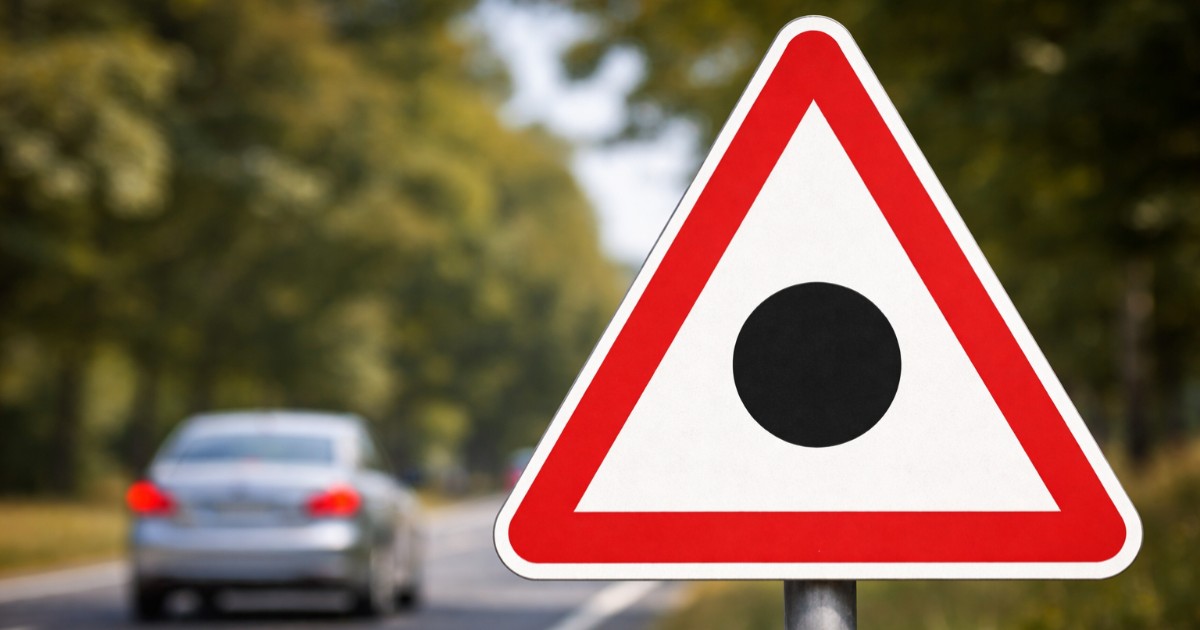 Imaginea prezintă un indicator rutier de avertizare triunghiular cu margine roșie și un punct negru în centru, amplasat pe marginea unui drum pe care circulă o mașină.