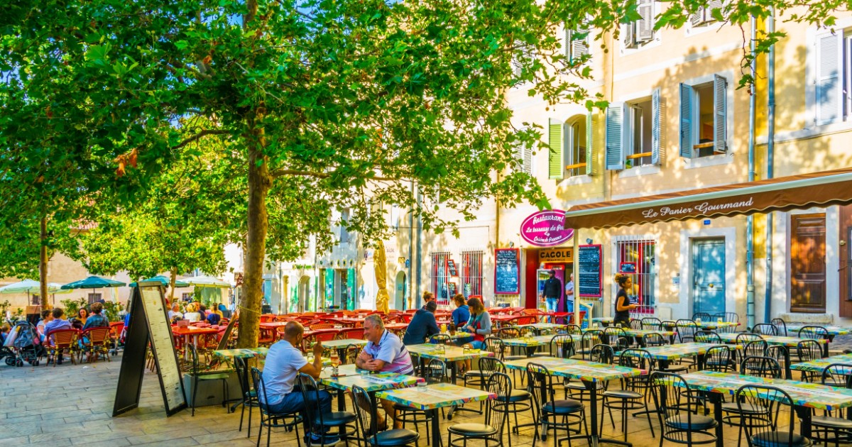 Imaginea surprinde o terasă animată a unui restaurant, probabil în Marsilia, Franța, cu mese și scaune negre, amplasată pe o piață pavată, umbrită de copaci verzi. Fațada clădirii din spate este de culoare galben pal, având obloane albastre și o marchiză maro pe care scrie "Le Panier Gourmand" (Panierul Gurmand).