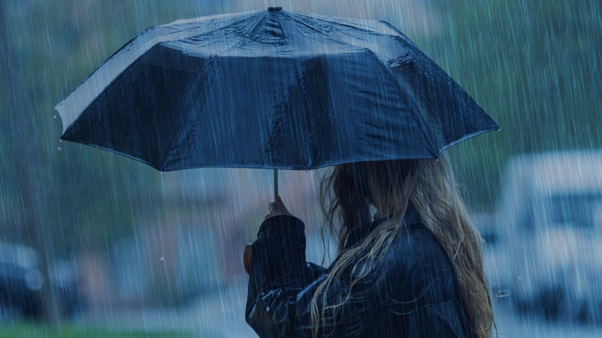 O persoană cu părul lung și blond, privită din lateral, care ține o umbrelă neagră deschisă pe o ploaie torențială.