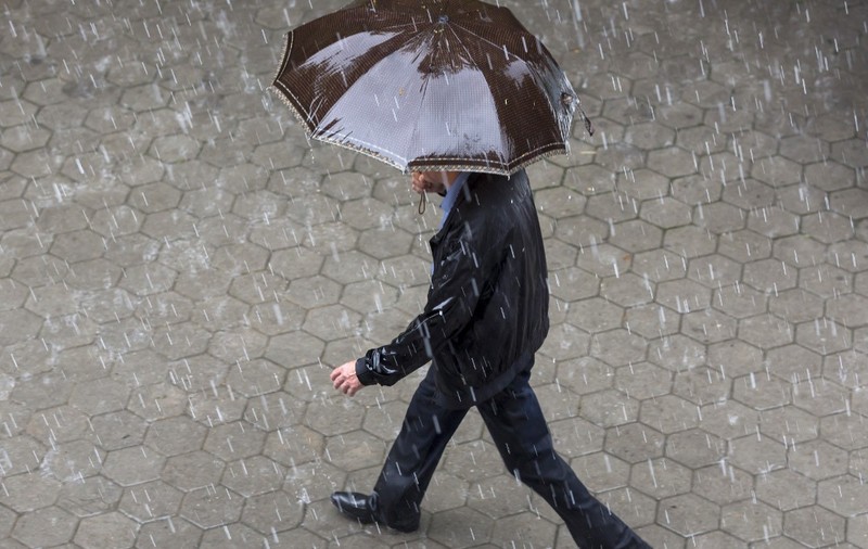 Un bărbat îmbrăcat într-o geacă neagră și pantaloni închiși la culoare, mergând pe un trotuar pavat în timp ce ține o umbrelă maro deasupra capului, pe o ploaie torențială.