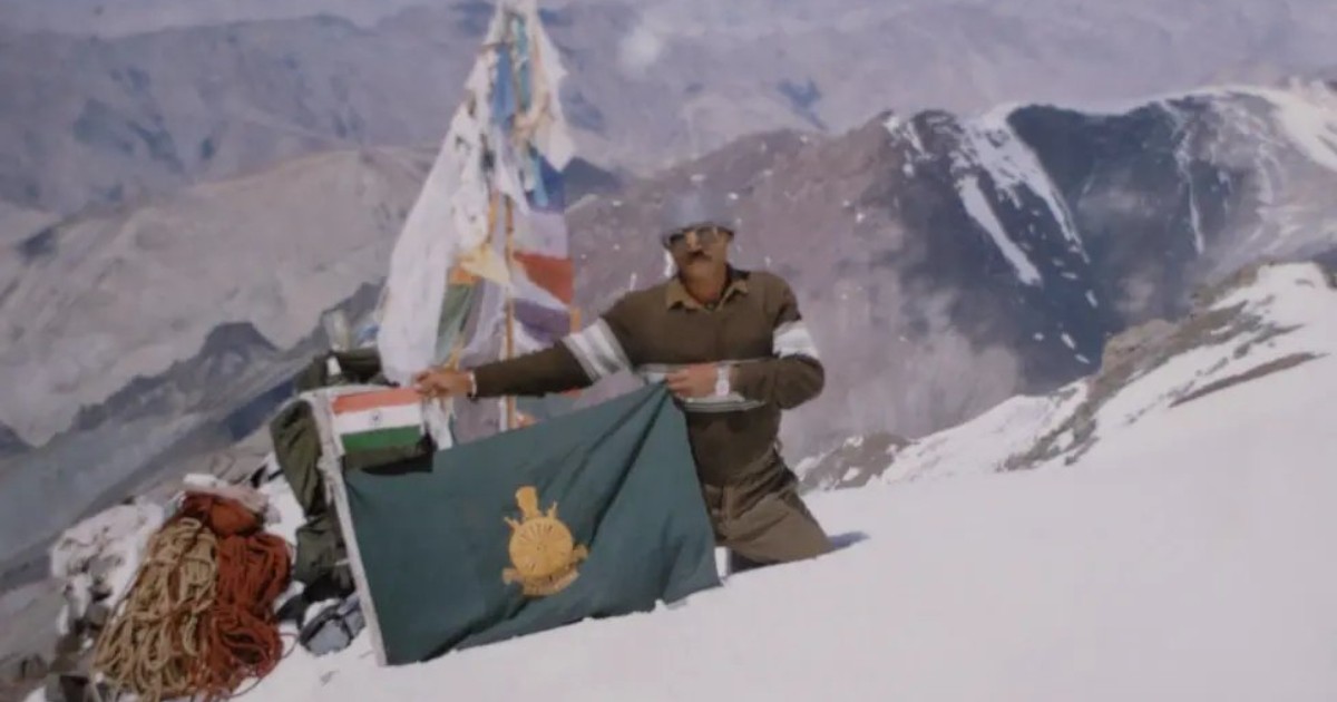 Imaginea prezintă un militar indian aflat într-o zonă montană acoperită de zăpadă, lângă steagurile Indiei și ale unei unități militare. În fundal se văd vârfuri muntoase înalte, iar în jurul său sunt echipamente de alpinism și corzi.