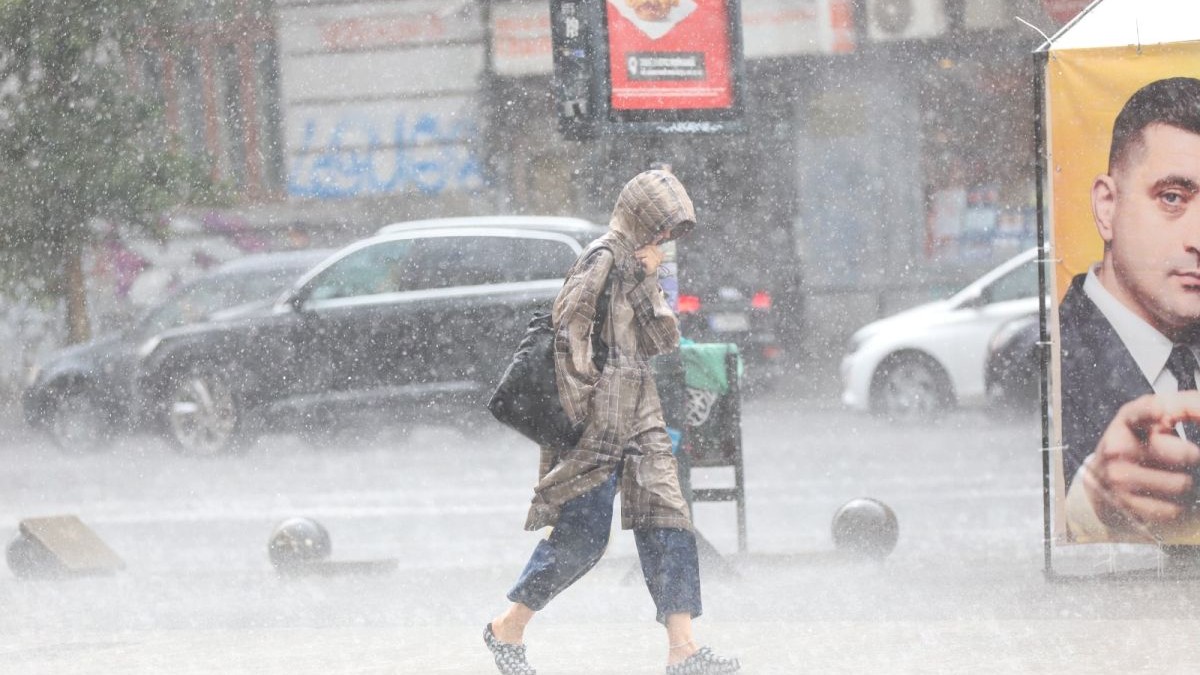 O femeie surprinsă de vremea rea în București merge pe stradă, se protejează de ploaie cu gluga și trece pe lângă un afiș electoral. Foto: Libertatea/Dumitru Angelescu
