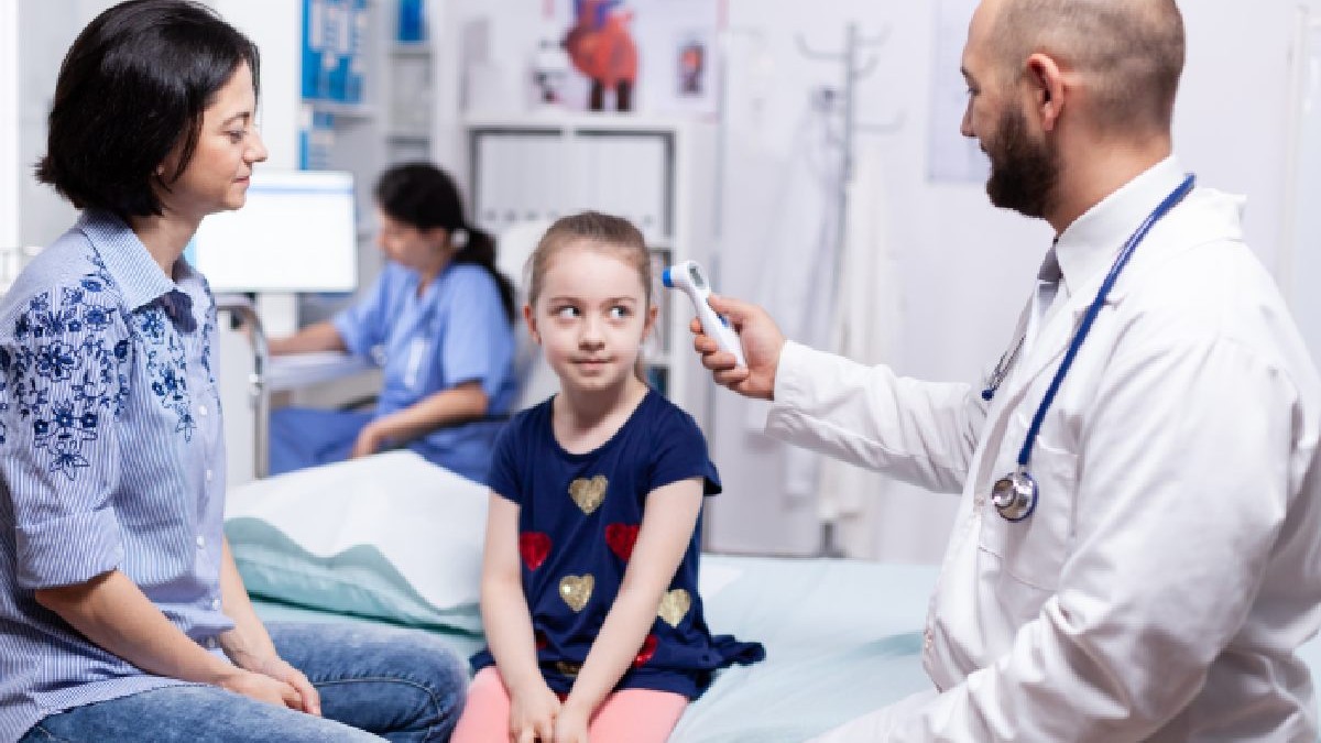 Un medic oncohematolog ia temperatura unei fetite care stă pe un pat de spital langa mama ei