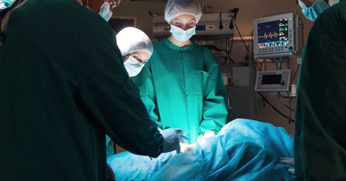 Medici chirurgi, imbracati in costume verzi si cu bonete pe cap, in sala de operatie, aplecati asupra unui pacient