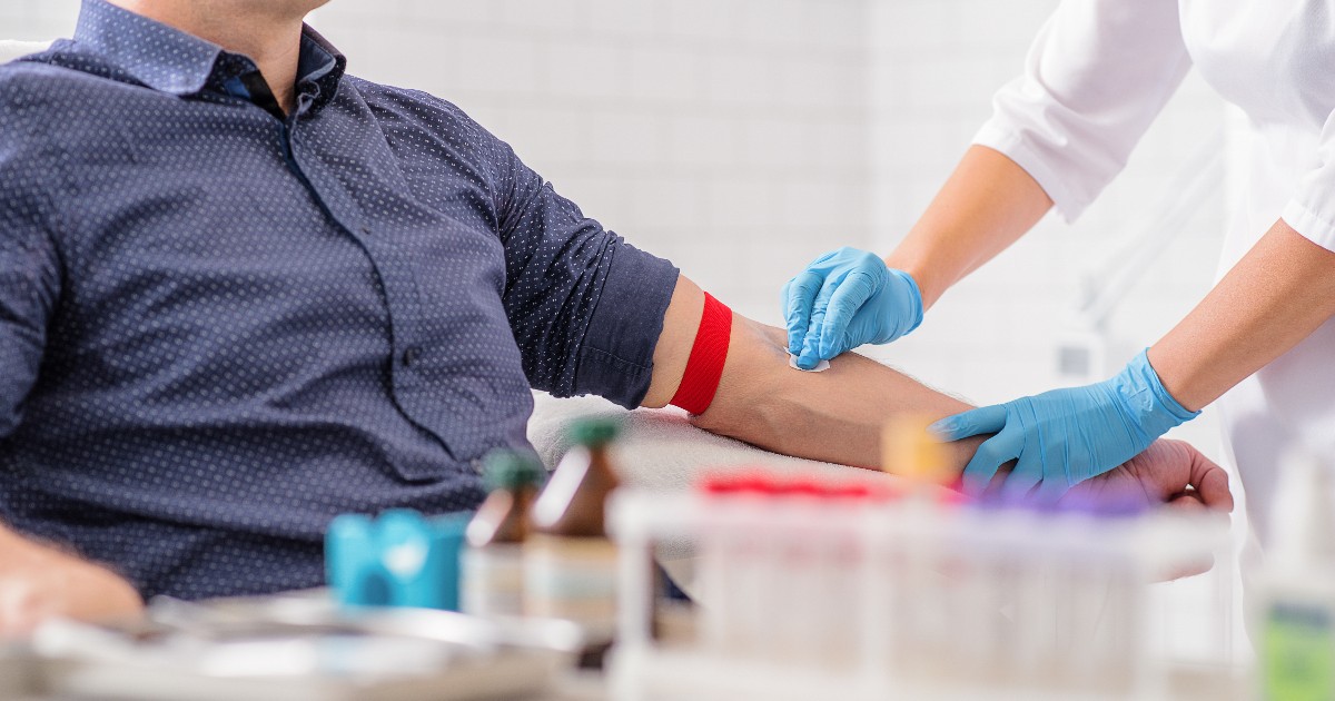 Imaginea prezintă un bărbat așezat pe un scaun, având o bandă roșie legată de brațul stâng, în timp ce o asistentă medicală, purtând mănuși albastre de protecție, îi dezinfectează cotul. Scena sugerează pregătirea pentru o procedură medicală, cel mai probabil o recoltare de sânge (analize).
