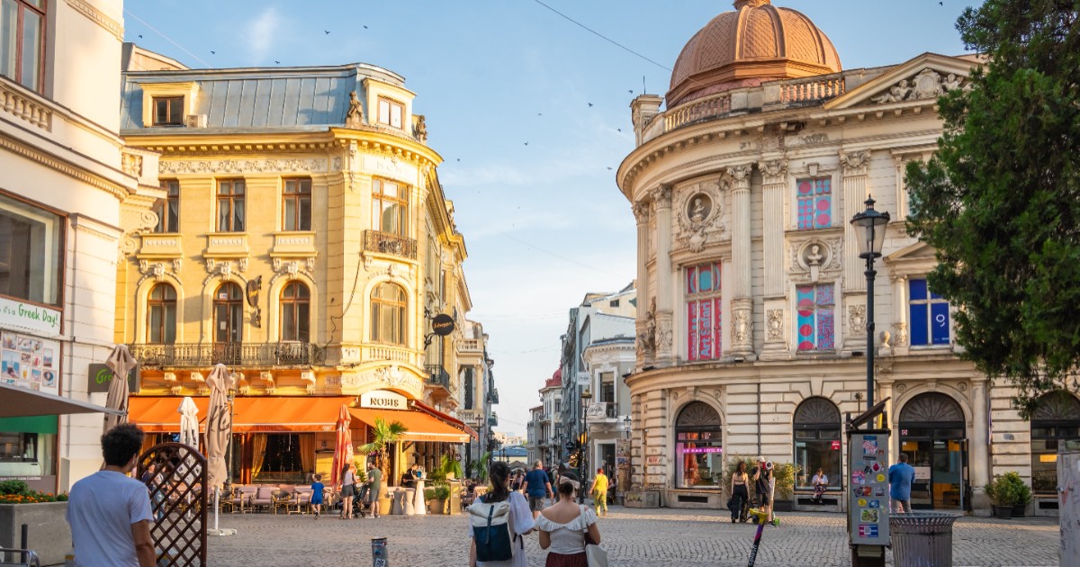 Turiștii plimbânduse prin Centrul Vechi din București, soarele bate pe clădirile istorice, în timp ce terasele sunt pline de clienți