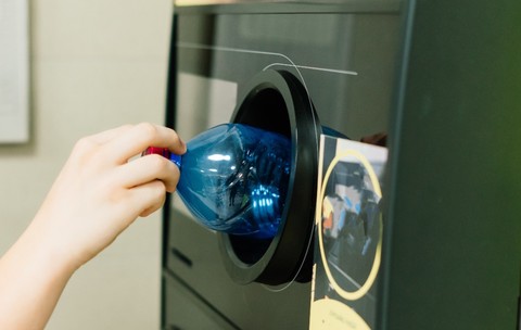 O mână care introduce o sticlă din plastic într-un aparat automat pentru colectarea și reciclarea ambalajelor.