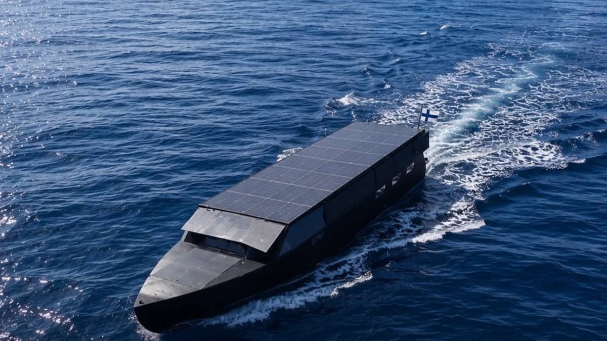 Iahtul solar Helios 11 navighează pe mare, cu acoperișul acoperit aproape integral de panouri fotovoltaice și steagul Finlandei la pupa.